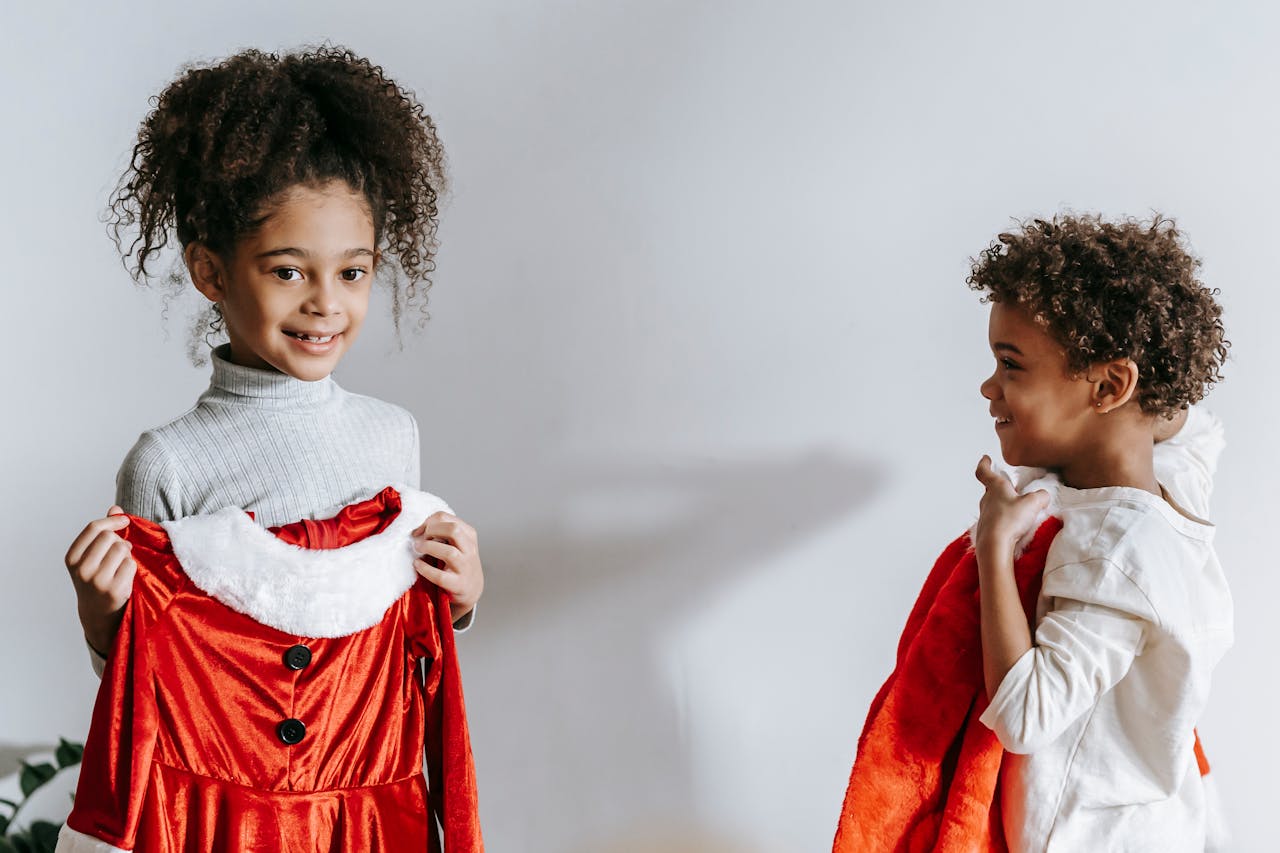 about-img Cheerful African American siblings with red Santa Claus outfits in hands standing in light room at home during Christmas holiday