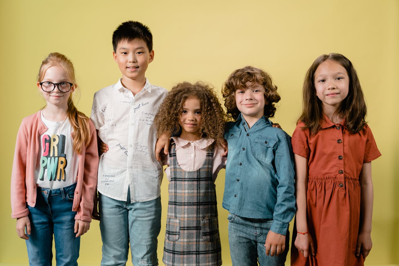 about-02 Five diverse children smiling against a yellow background in a studio setting.